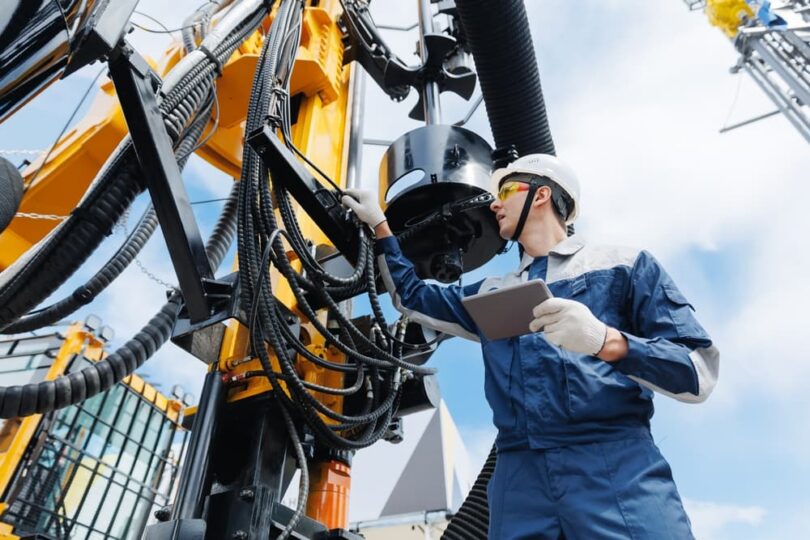 Oilfield technician inspecting drilling equipment while holding a tablet and wearing protective gear